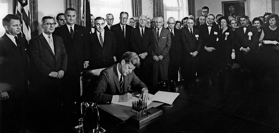 Black and white photo of President Kennedy sitting at desk and signing document. A group of men and women in suits stands behind him and looks on.