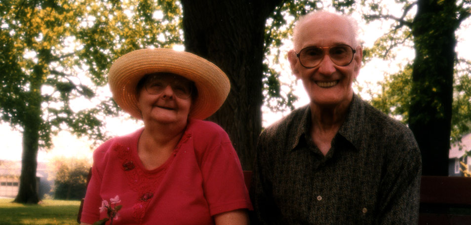 Photo of an older couple smiling and sitting on a park bench. The woman is wearing a sun hat